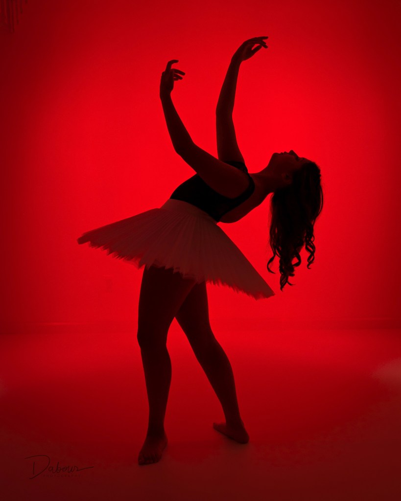 Ballet dancer silhouetted against red background with arms extended and head tilted back.