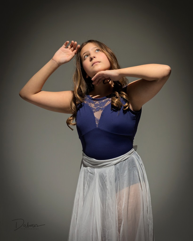 Dancer in navy leotard and sheer skirt posing with raised arms under studio lighting.