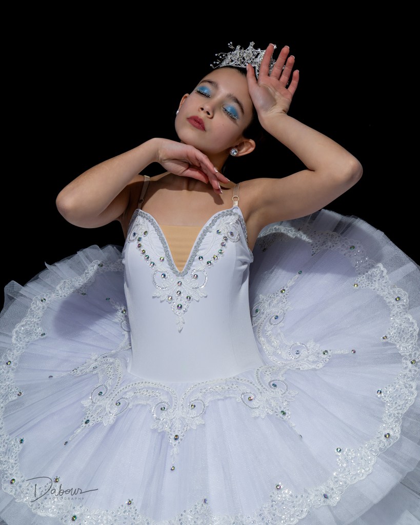 dancer in white with a large tutu on and crown with eyes closed feeling the light above her face