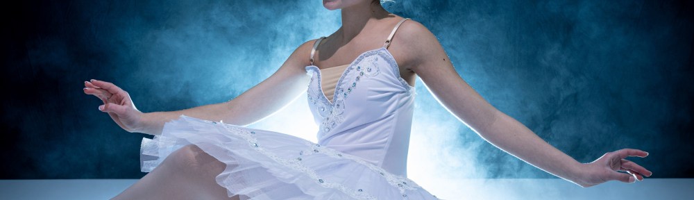 Ballet dancer Lucy seated in white tutu with tiara, arms extended gracefully, against dramatic blue mist.