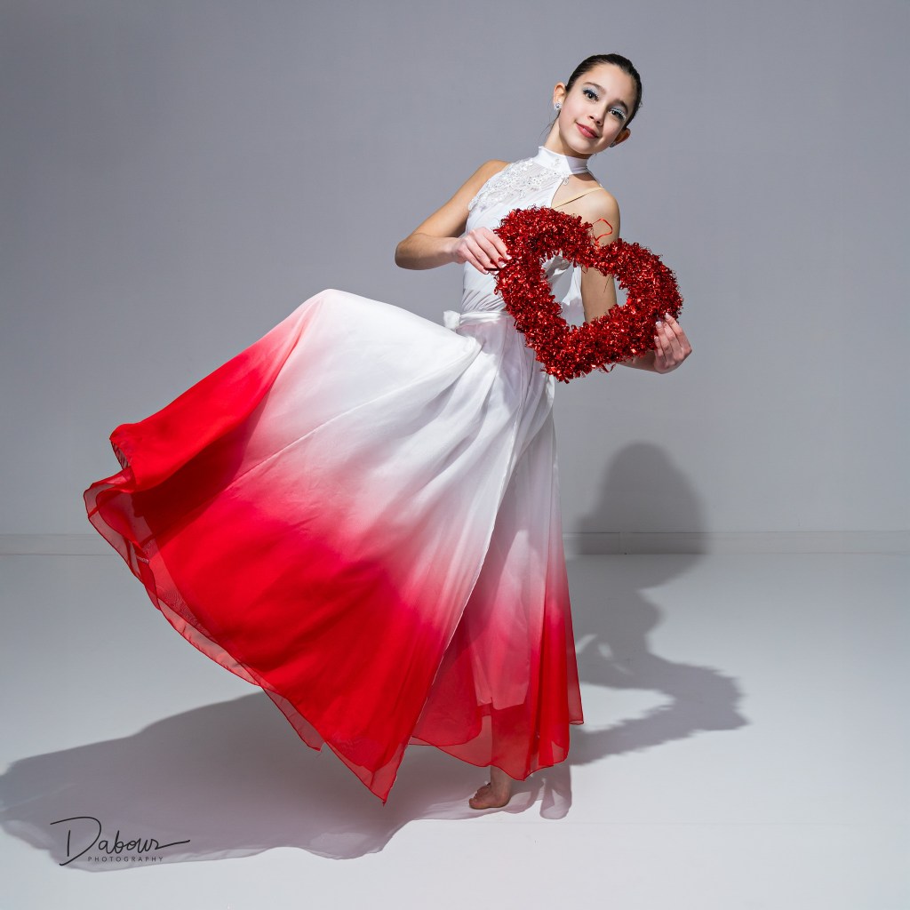Young dancer in a white and red flowing dress holding a red heart prop in a Valentine themed studio portrait.