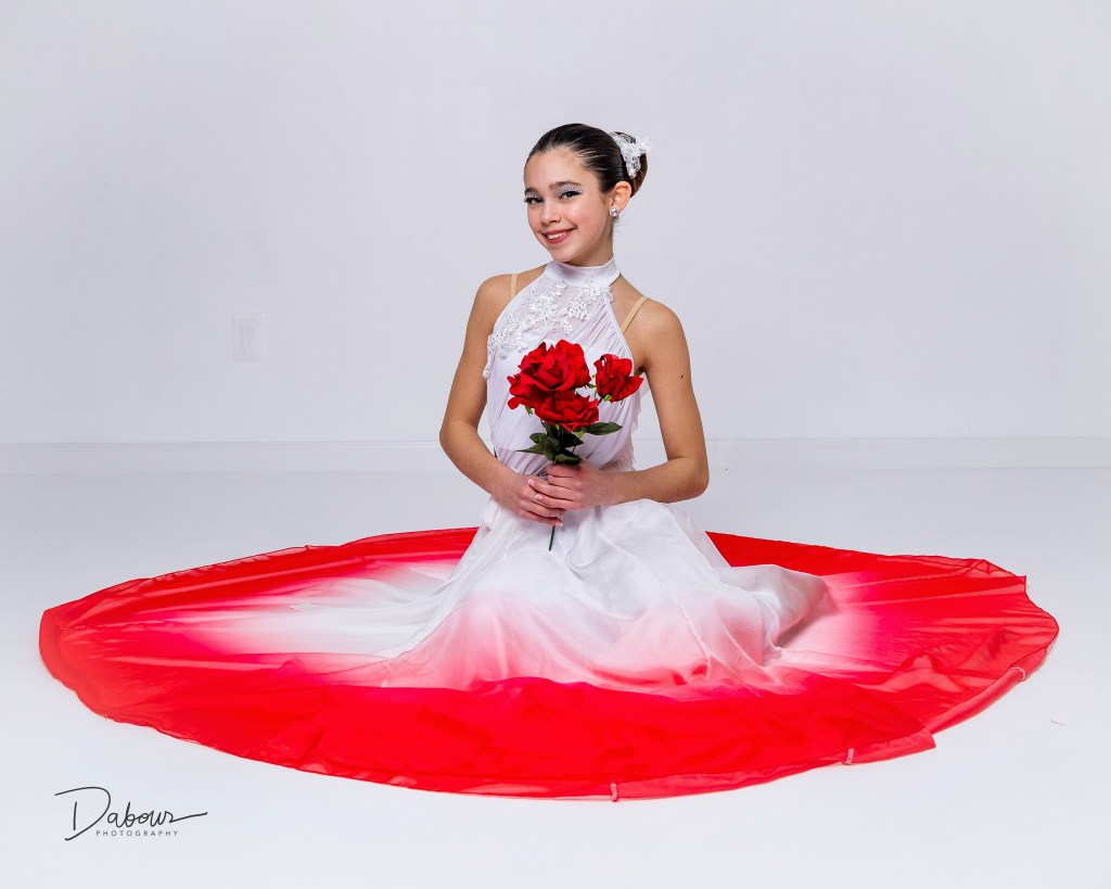 Young dancer seated on the studio floor holding red roses with a red and white flowing skirt arranged around her.