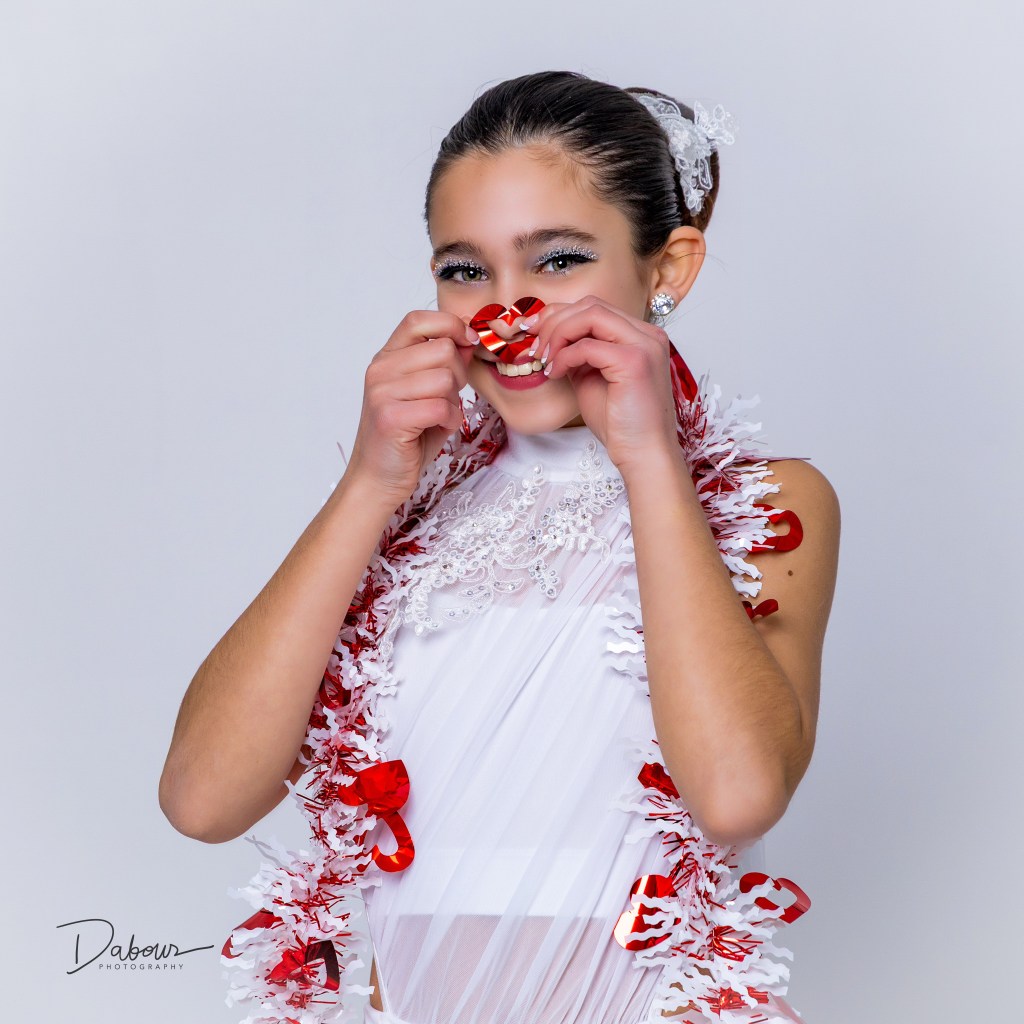 Smiling dancer holding a small heart near her face while wearing Valentine themed garland in a studio portrait.