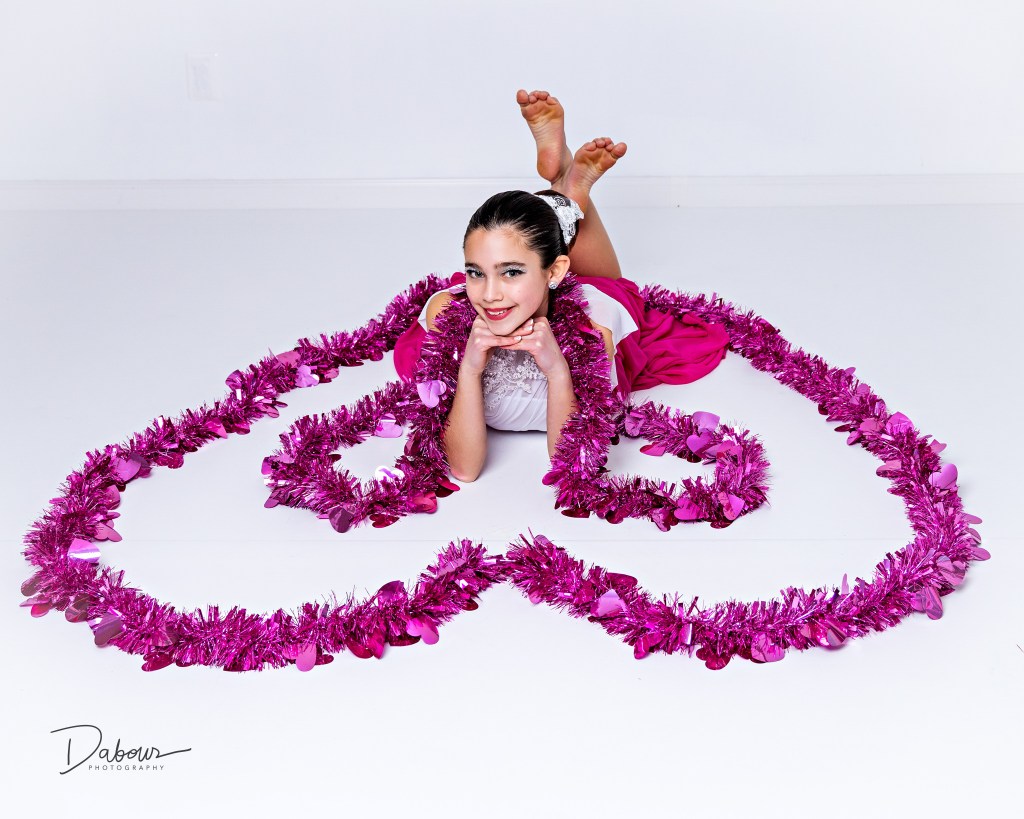Young dancer lying on the studio floor surrounded by pink heart shaped garland arranged in a Valentine design.
