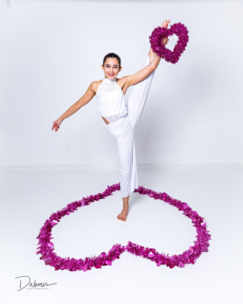 Dancer performing a high leg extension while holding a pink heart prop during a Valentine themed studio photo session.
