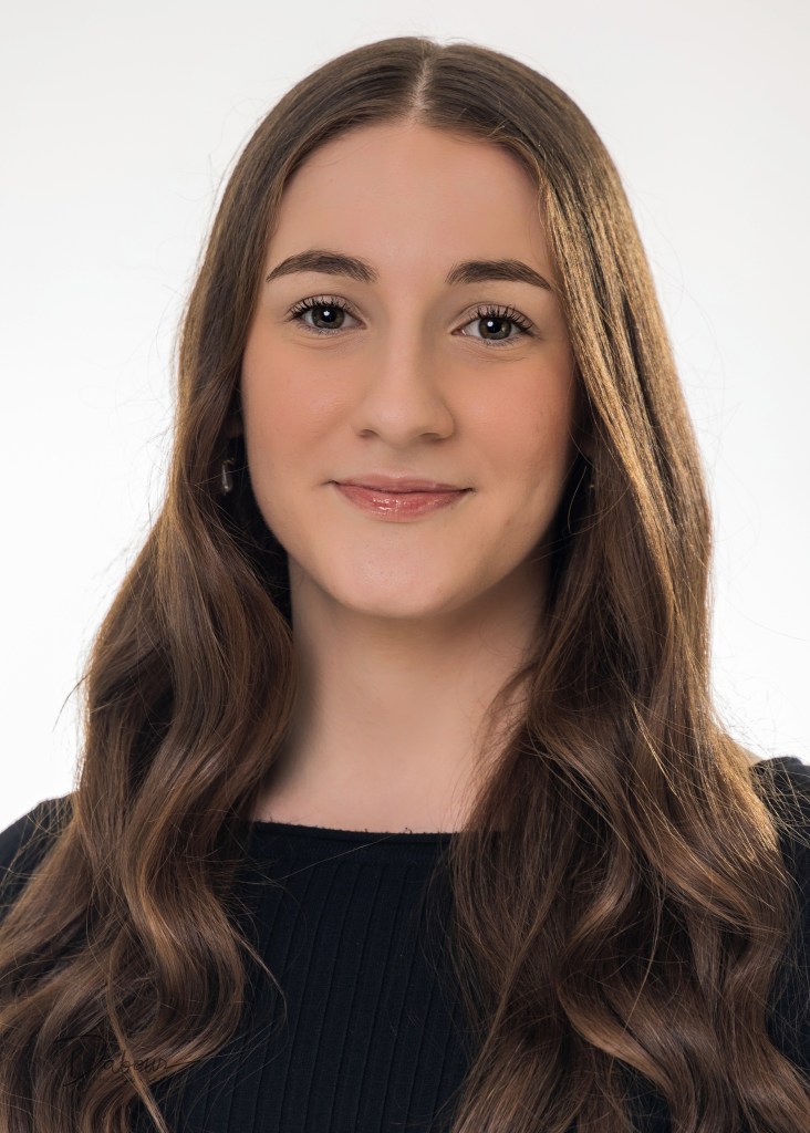 A professional headshot of Riley, a young woman with long brown hair, smiling warmly. She is wearing a dark top against a neutral, light-colored studio background with soft lighting.