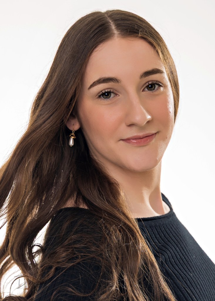 A professional headshot of Riley, a young woman with long dark hair, looking directly at the camera with a neutral expression. She is wearing a dark top against a soft-focus, light studio background.