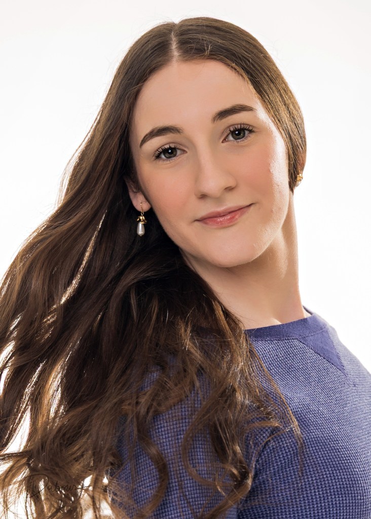 A professional headshot of Riley, a young woman with long dark hair, looking directly at the camera with a gentle, confident expression. She is wearing a dark top against a soft, light-colored studio background.