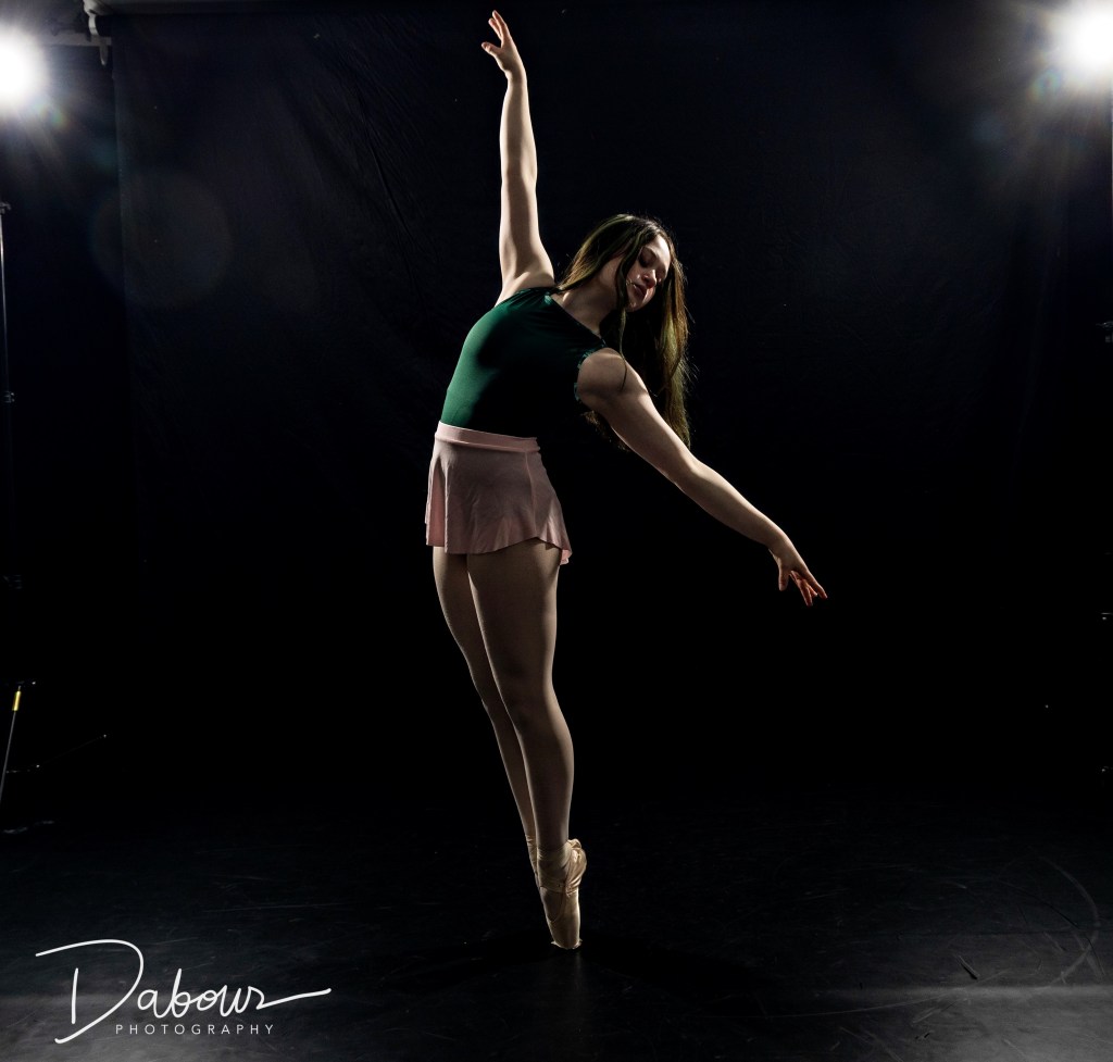 A dance ambassador performing a dramatic high kick in a dark studio corner, illuminated by sharp strobe lighting that creates high-contrast shadows against the black walls.