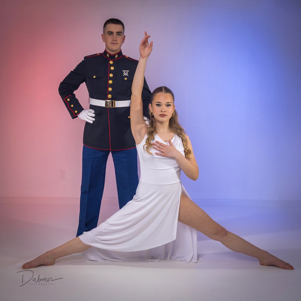 A US Marine in a Dress Blue uniform stands proudly next to a ballet dancer in a white dress and pointe shoes in a professional studio setting.