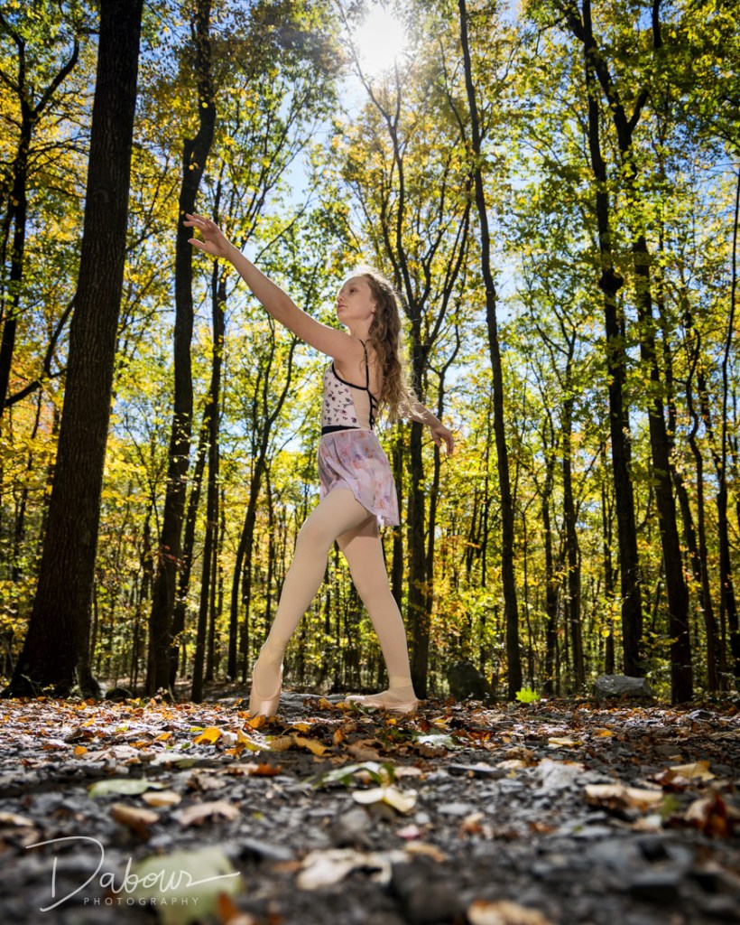 A young ballet dancer, Lily, performing a high leg extension (developpé) while standing on a forest path at Columcille Megalith Park. She is wearing a purple dance skirt and floral leotard, surrounded by tall trees and stone monuments.