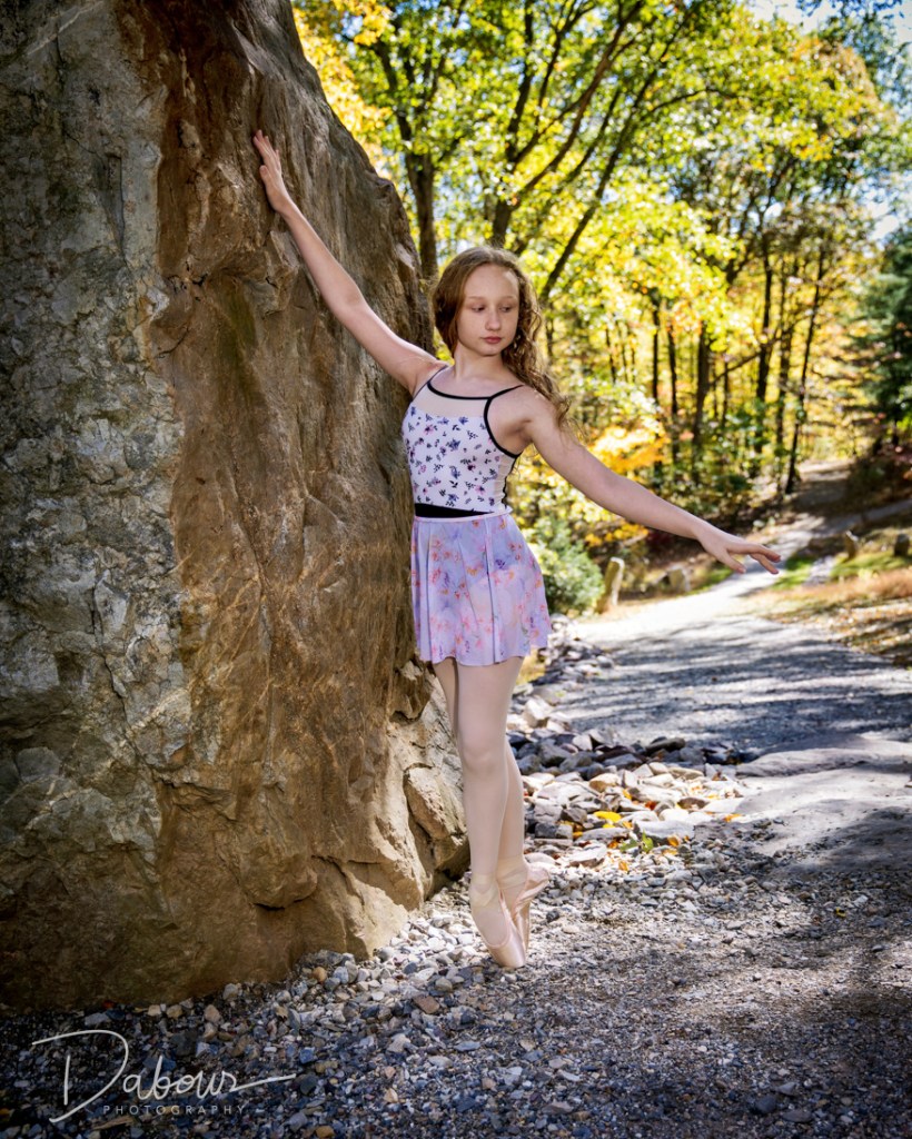 A young ballet dancer, Lily, posing against a towering, narrow standing stone at Columcille Megalith Park. She is wearing a floral patterned leotard and a light purple skirt, standing on pointe with one arm reaching up the textured rock face and the other extended gracefully toward the forest path.