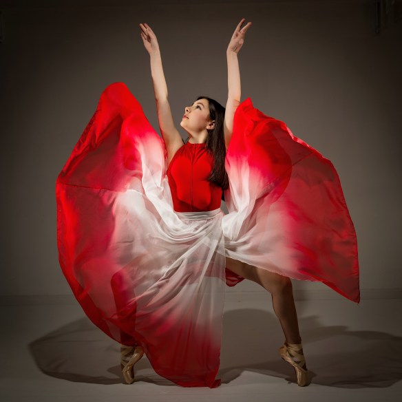 A female dancer in a white leotard and flowing red skirt performing an expressive, mid-motion pose against a professional gray studio backdrop.