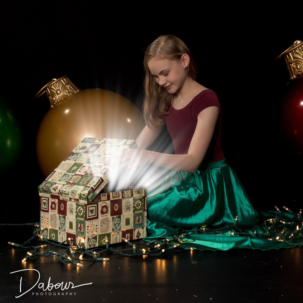 A young dancer, Sydney, in a maroon velvet top and emerald green satin skirt, kneeling on a reflective floor. She is looking down with a smile into a decorative holiday gift box that is emitting a bright, magical white glow, surrounded by Christmas ornaments and fairy lights.