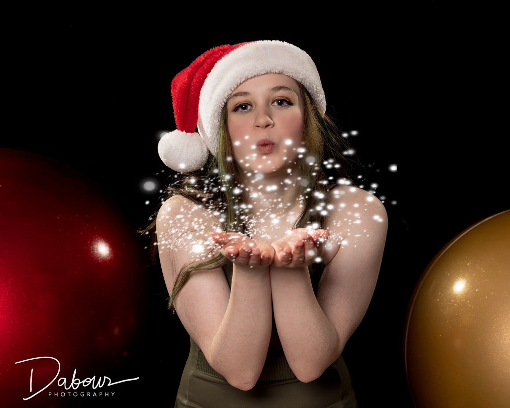 A dancer in a red top and Santa hat posing against a dark background with blurred, glowing red and white holiday bokeh lights.