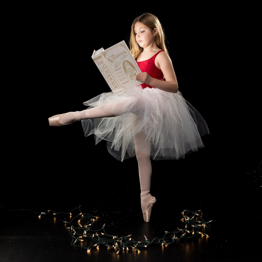 Young ballerina Vivi Taggart posing in front of a glowing Christmas tree and holiday lights at West Jersey Youth Ballet.