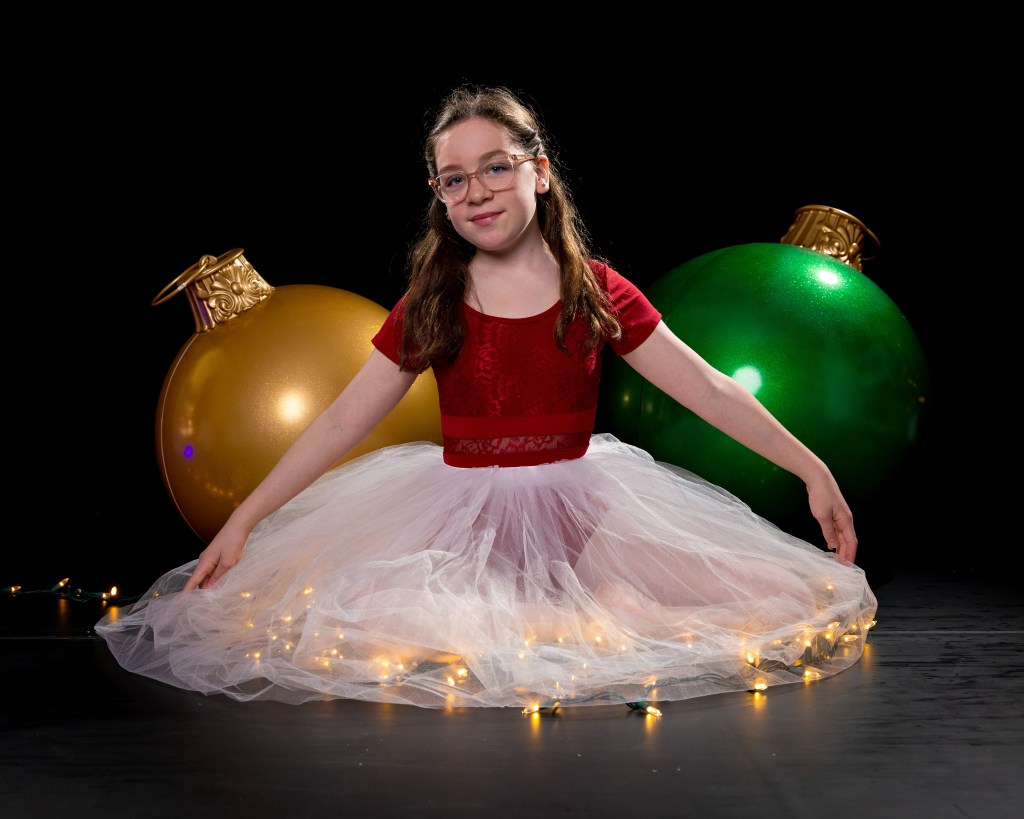 Young ballerina Olivia Klein in a red and white holiday dance costume, sitting gracefully on the floor surrounded by glowing gold and green Christmas ornaments and festive studio lights.