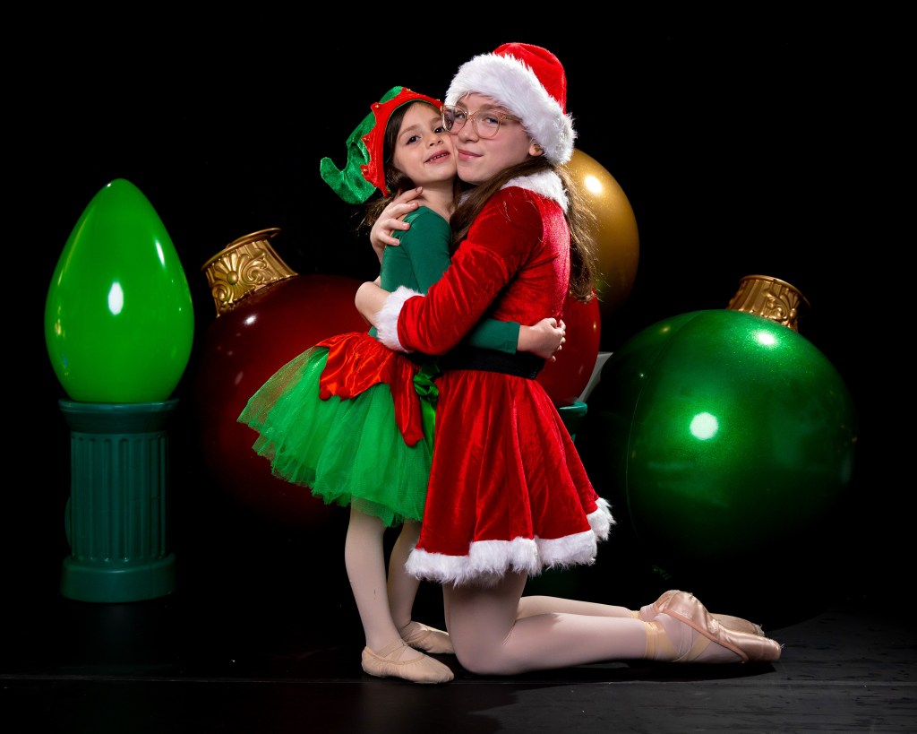 Two young sisters, Olivia and Brielle Klein, dressed in matching red and white holiday dance outfits, hugging and smiling together in a festive Christmas-themed studio set.