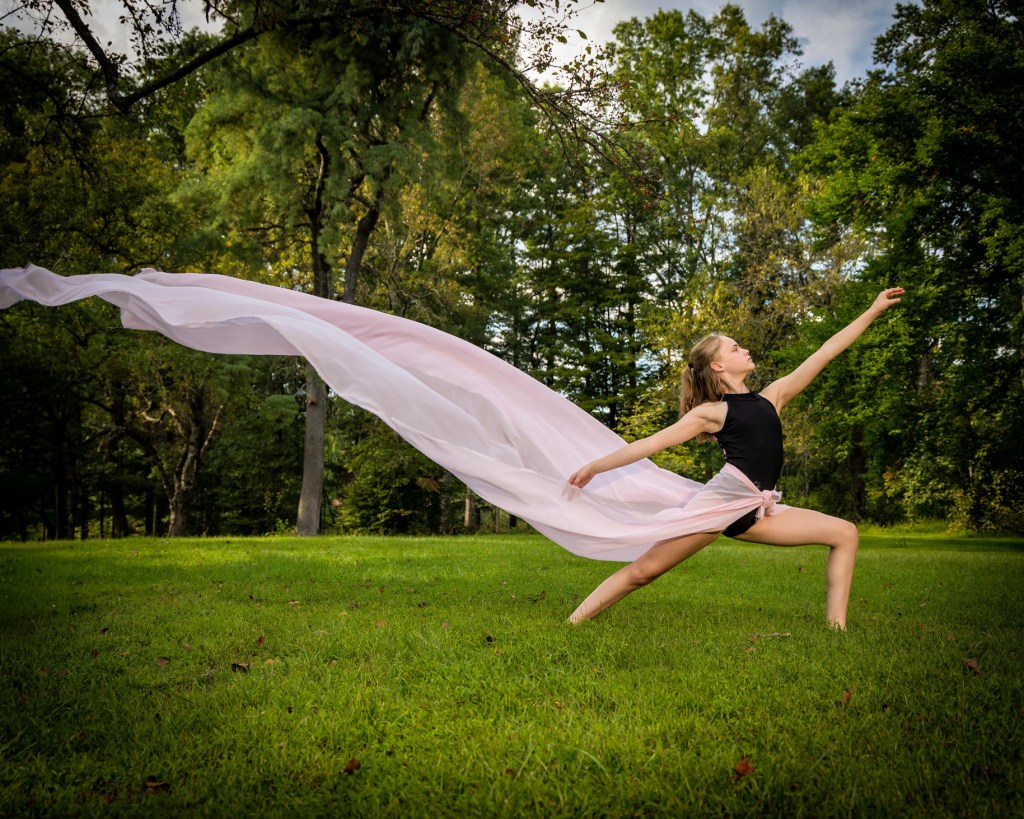 A young dancer, Sydney, in a black leotard performing a deep lunge (second position) on a green grass field. A long, translucent light pink scarf is blowing dramatically in the wind behind her, framed by lush green trees and a bright sky.