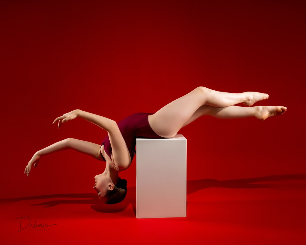 Artistic dance portrait of Lily kneeling on a white studio block against a deep red background. She is wearing a maroon velvet leotard and looking toward the camera with a focused, dramatic expression.