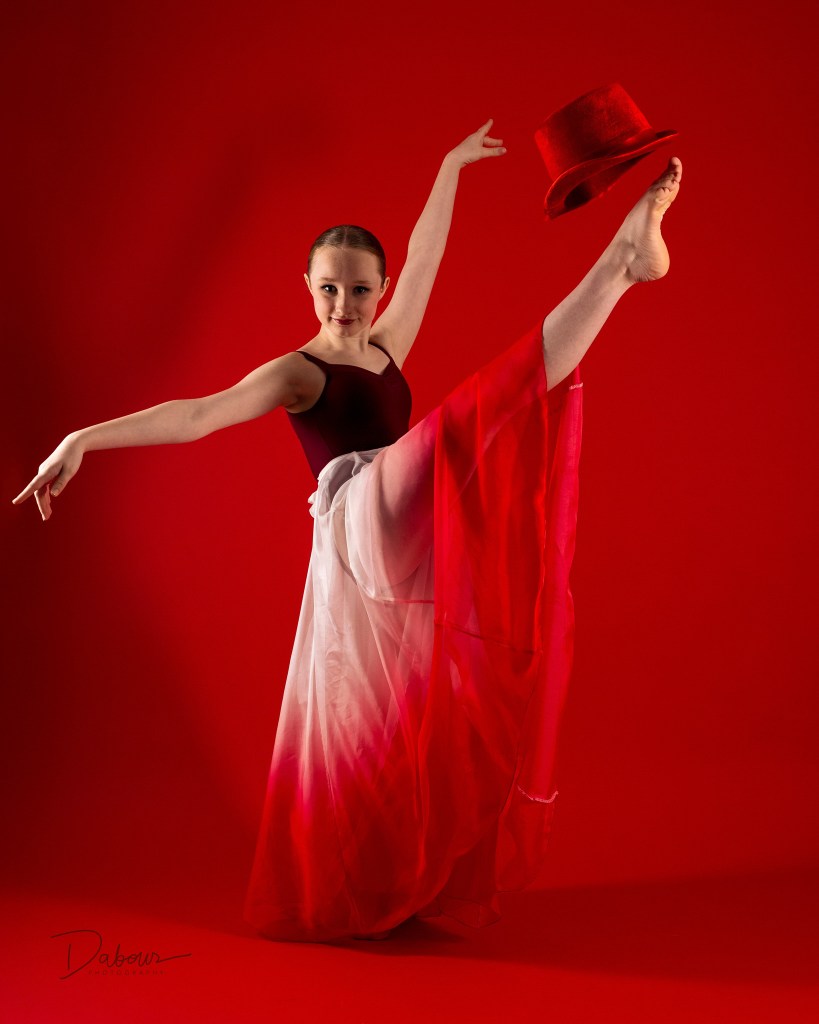 Artistic dance portrait of Lily kneeling on a white studio block against a deep red background. She is wearing a maroon velvet leotard and looking toward the camera with a focused, dramatic expression.