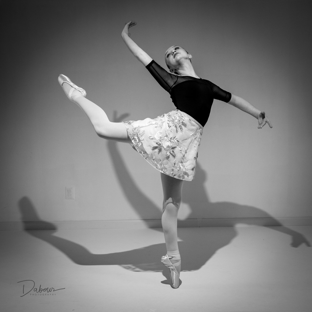 A high-fashion dance portrait of Lily leaning back against a white block on a reflective studio floor. The monochromatic red background highlights her maroon leotard and athletic silhouette.