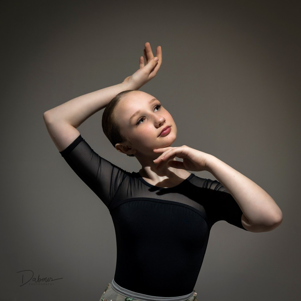 A high-fashion dance portrait of Lily leaning back against a white block on a reflective studio floor. The monochromatic red background highlights her maroon leotard and athletic silhouette.