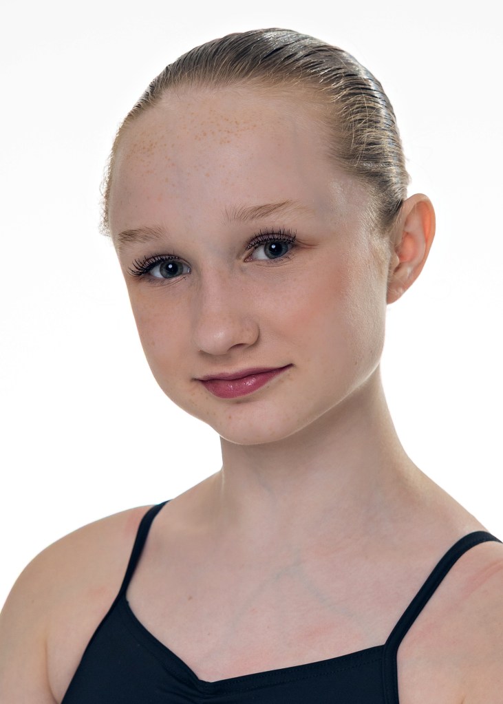 A professional close-up headshot of Lily, a young ballet dancer, with a soft, natural smile. She has light hair and is wearing a dark dance leotard against a simple studio background.