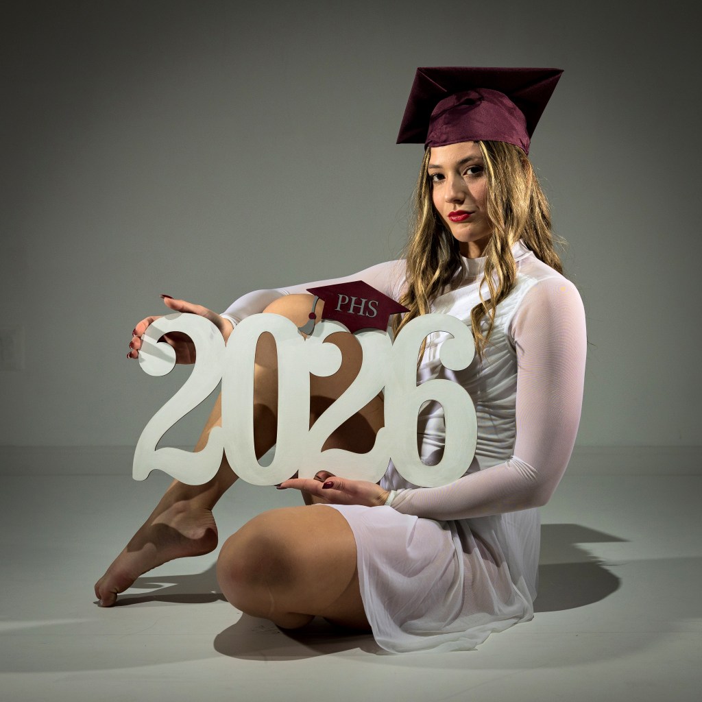 Senior portrait of Mia wearing a graduation cap and gown, sitting behind large "2026" props in a professional studio setting.