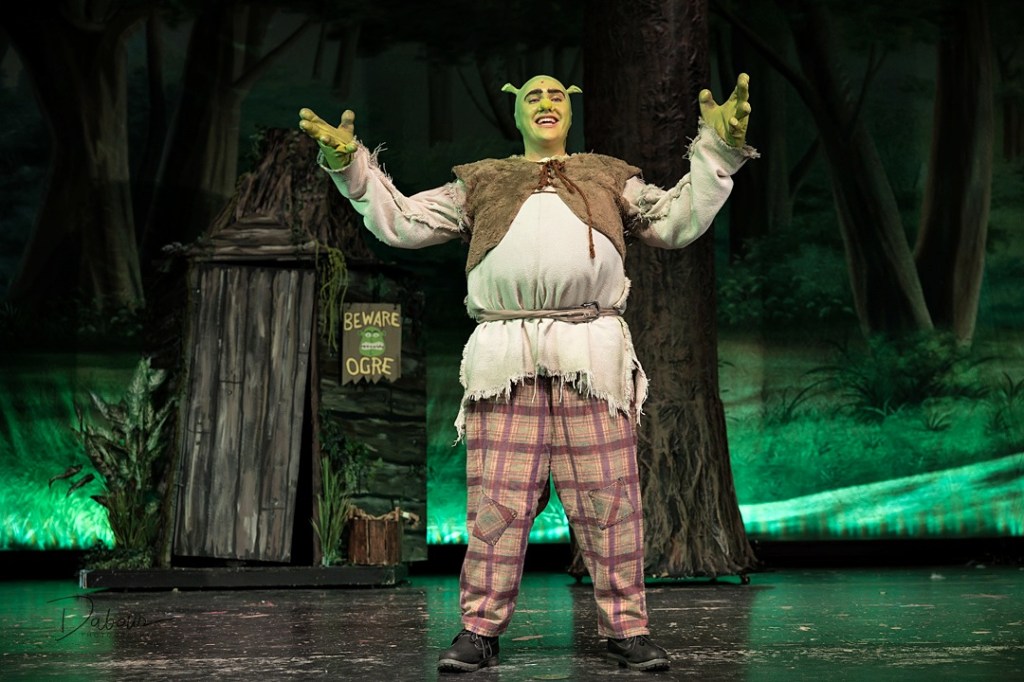 A stage production photo of a student performer dressed as Shrek from "Shrek the Musical." The actor is in full green ogre makeup and costume, standing in a swamp-themed set with a "Beware Ogre" sign and large trees in the background. He is posing with his arms out and an expressive facial expression under theatrical green lighting.