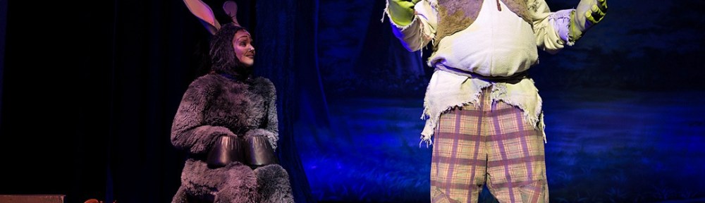 A stage production photo from "Shrek the Musical" at North Hunterdon High School. Shrek, in his green ogre costume and plaid pants, stands next to Donkey, who is wearing a grey fur costume with long ears. They are performing in front of a forest backdrop under theatrical lighting.