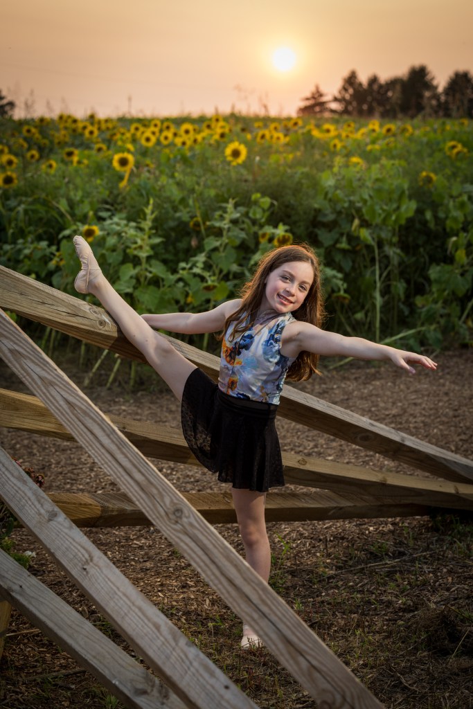 Ballerina Olivia Klein posing in a black leotard and floral skirt in the middle of a vibrant yellow sunflower field, reaching one arm up gracefully.