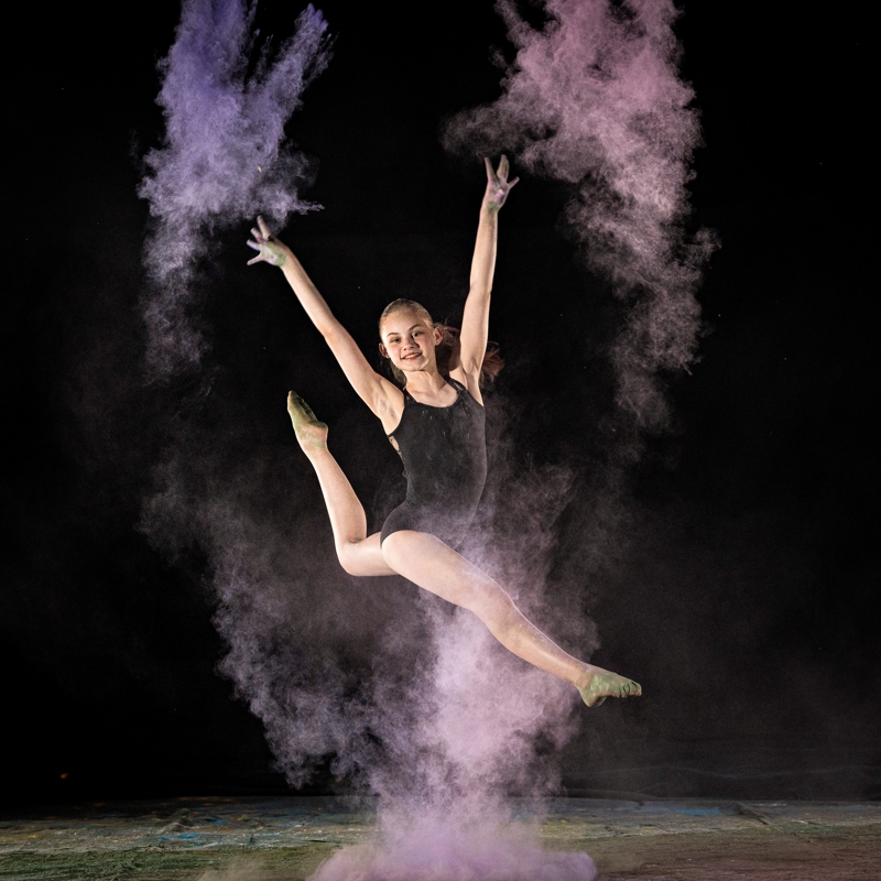A high-action dance portrait of Sydney performing a grand jeté (split jump) in mid-air. She is surrounded by a large, explosive cloud of purple powder that follows her movement against a solid black background.