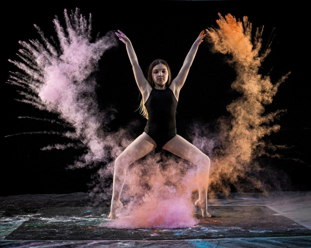Dynamic action shot of a dancer performing a jump with white flour or powder exploding around her for a high-contrast creative effect.