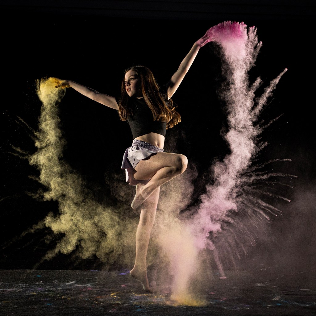A creative dance portrait of a ballerina in a dark leotard, with white powder forming a glowing heart shape around her as she poses in a studio.