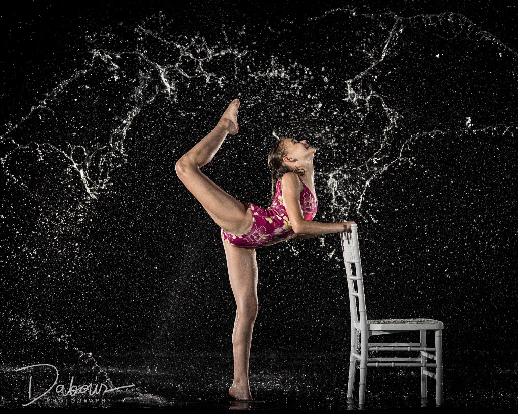 A dancer, Sydney, in a purple floral leotard, performing a back-bending pose while leaning against a white wooden chair in a rain machine. Water is splashing and frozen in droplets around her against a dark background.
