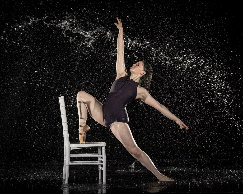 A dance ambassador posing dynamically under falling water in a rain machine studio session, with droplets frozen in mid-air by strobe lighting.