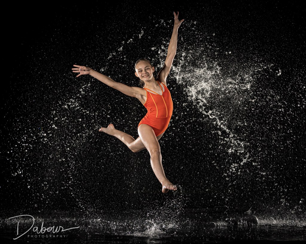 A high-action dance portrait of Sydney in a rain machine. She is performing a vertical leg extension (developpé) while wearing a bright orange leotard. Water splashes and droplets are frozen in mid-air around her against a dark, dramatic background.