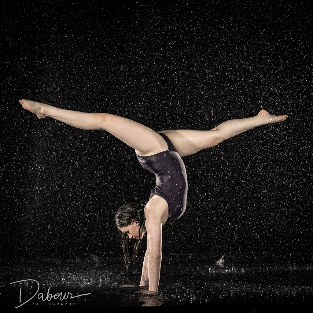 A dancer mid-motion during a rain machine photo session, featuring dramatic lighting and splashing water on a dark studio floor.