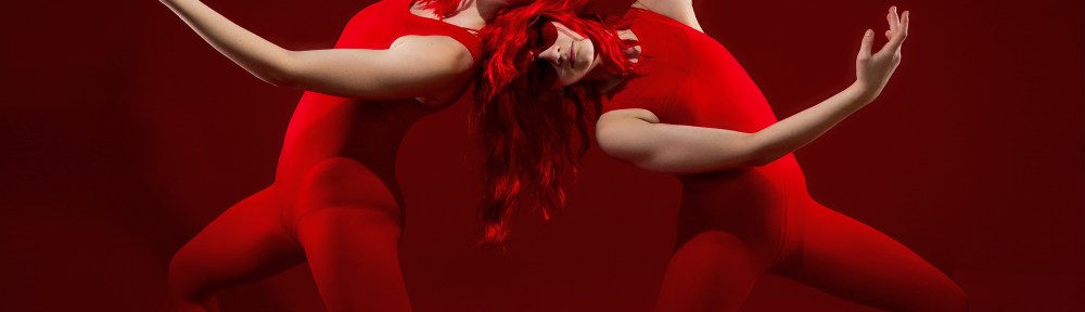 Two dance ambassadors in red wigs and bodysuits performing a symmetrical, artistic "V" shape pose against a solid red backdrop in a studio.