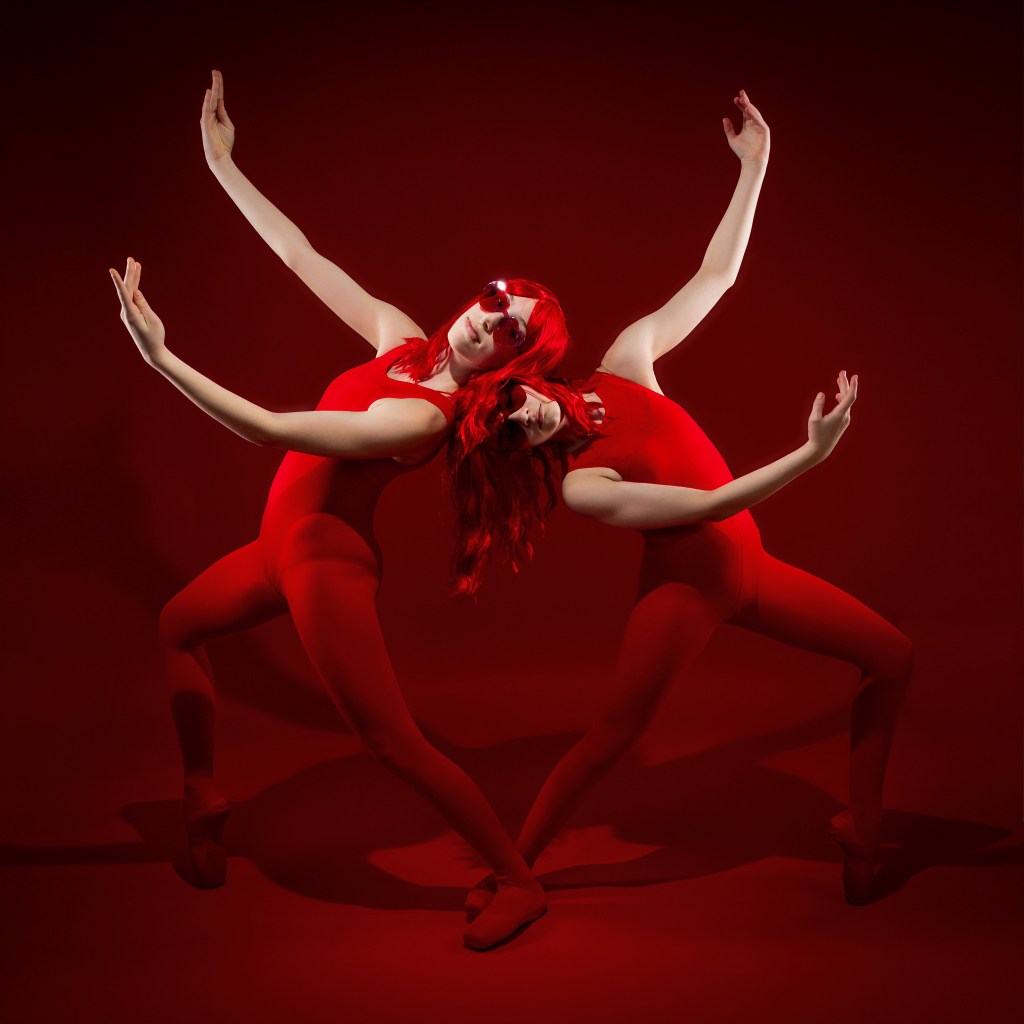 Ellery and Sydney posing together in red wigs and red bodysuits against a bold red backdrop during their Scarlet Sisters dance portrait session.