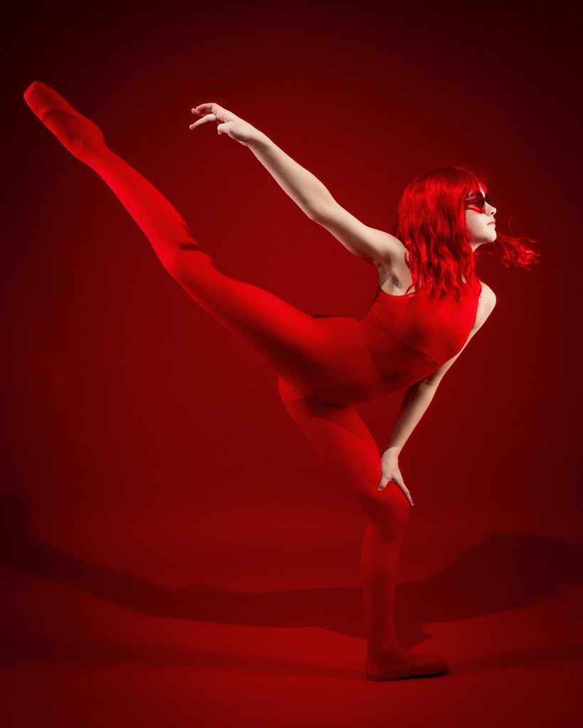 A high-energy dance portrait of Sydney in a monochromatic red theme. She is wearing a red bodysuit, red tights, and red sunglasses, performing a high leg extension (arabesque) while balanced on one leg against a solid red background.