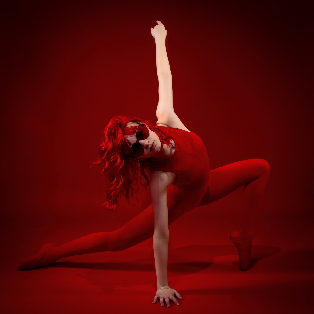 A dynamic dance portrait of Sydney in a monochromatic red theme. She is wearing a red bodysuit, red tights, and red sunglasses, posing in a deep lunge with one arm extended straight upward against a vibrant red background.