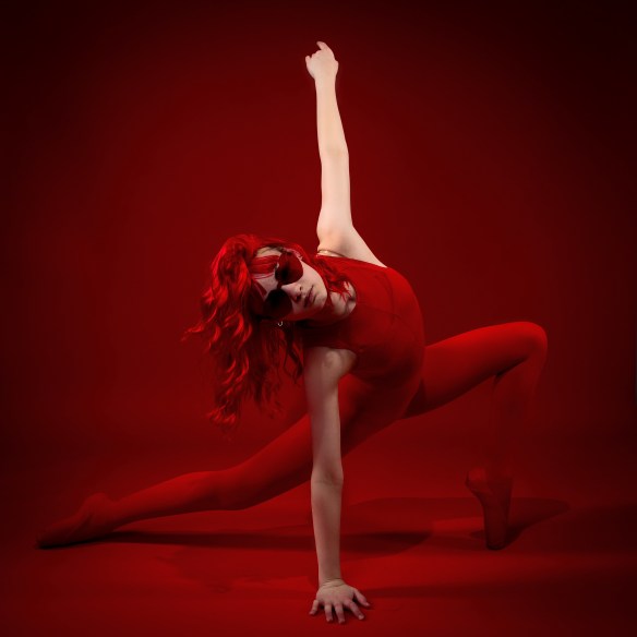A dynamic dance portrait of Sydney in a monochromatic red theme. She is wearing a red bodysuit, red tights, and red sunglasses, posing in a deep lunge with one arm extended straight upward against a vibrant red background.
