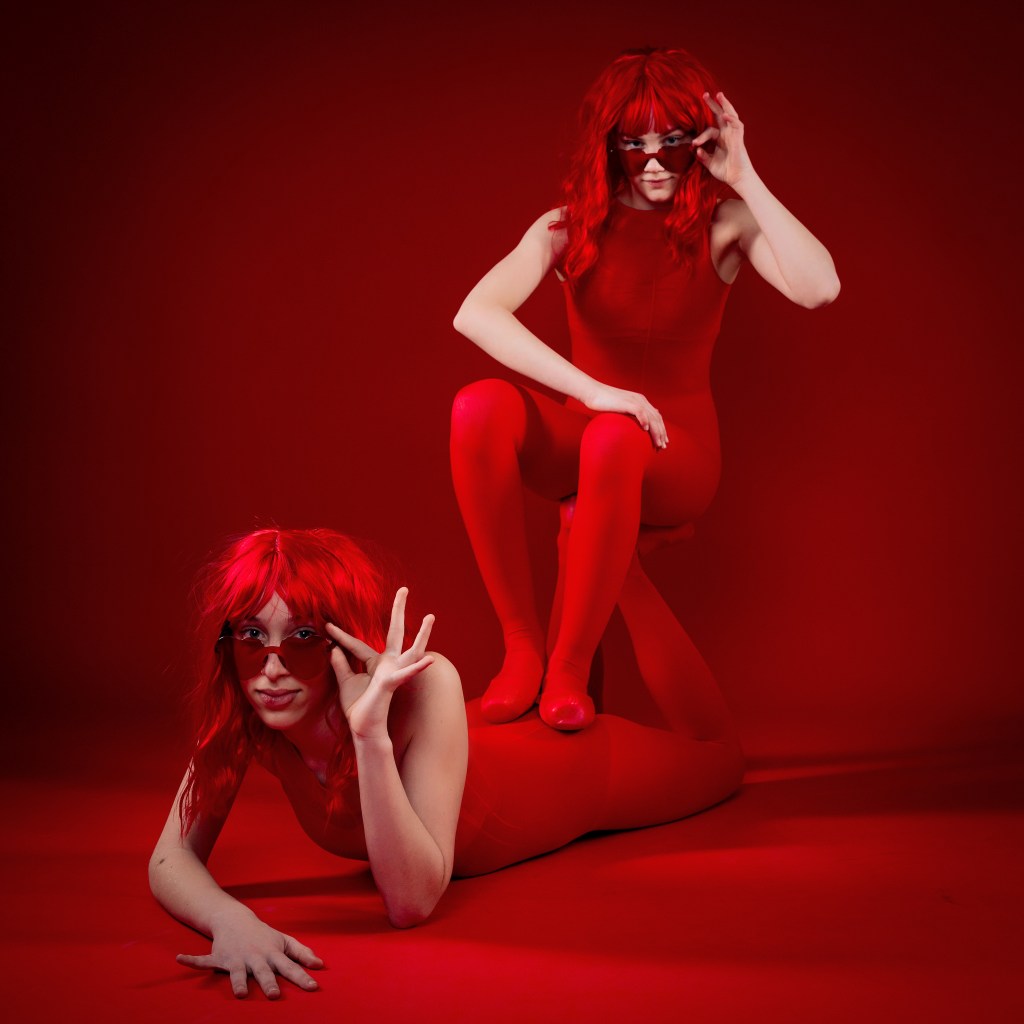 Ellery and Sydney posing together in red wigs and red bodysuits against a bold red backdrop during their Scarlet Sisters dance portrait session.