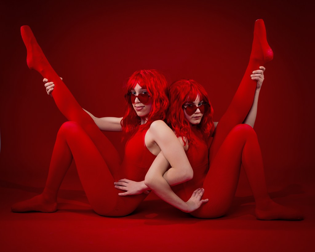 Ellery and Sydney posing together in red wigs and red bodysuits against a bold red backdrop during their Scarlet Sisters dance portrait session.