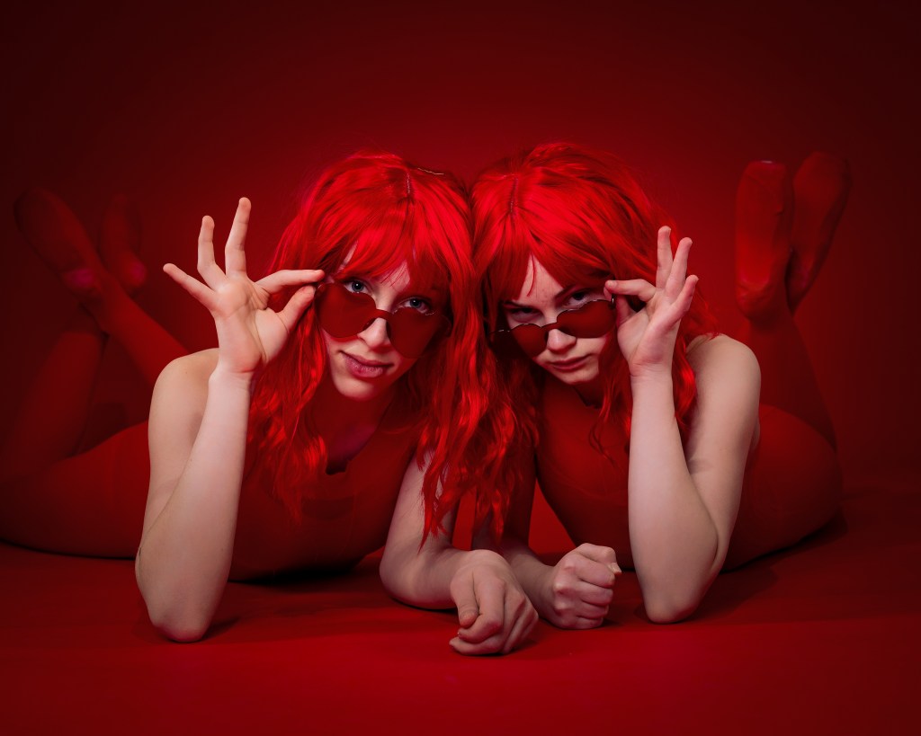 Ellery and Sydney posing together in red wigs and red bodysuits against a bold red backdrop during their Scarlet Sisters dance portrait session.