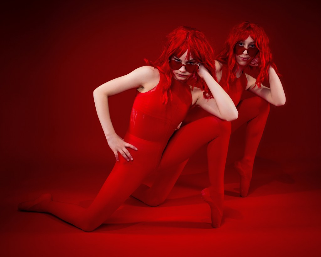 Ellery and Sydney posing together in red wigs and red bodysuits against a bold red backdrop during their Scarlet Sisters dance portrait session.
