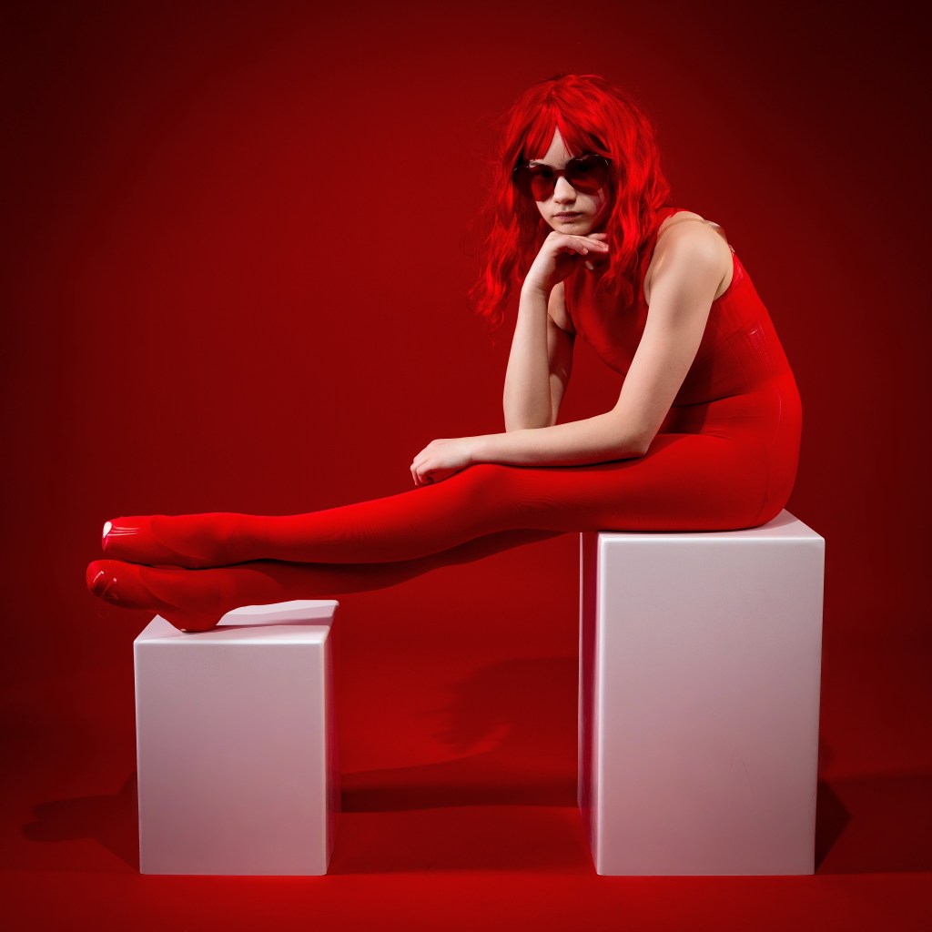 A high-fashion dance portrait of Sydney in a monochromatic red theme. She is wearing a red sleeveless bodysuit, red tights, and red sunglasses with her hair dyed bright red. She is sitting in a relaxed, thoughtful pose on white minimalist blocks against a deep red background.