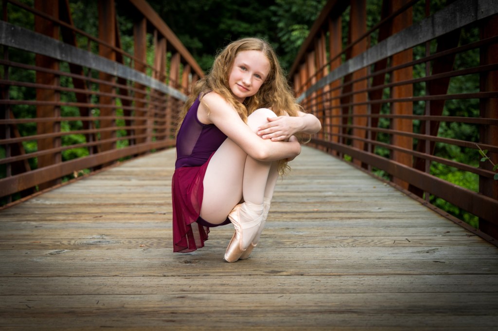 Lily performing a graceful ballet pose on a wooden bridge at the Easton Silk Mill. She is on pointe, wearing a maroon leotard, with one arm extended toward the industrial brick architecture in the background.
